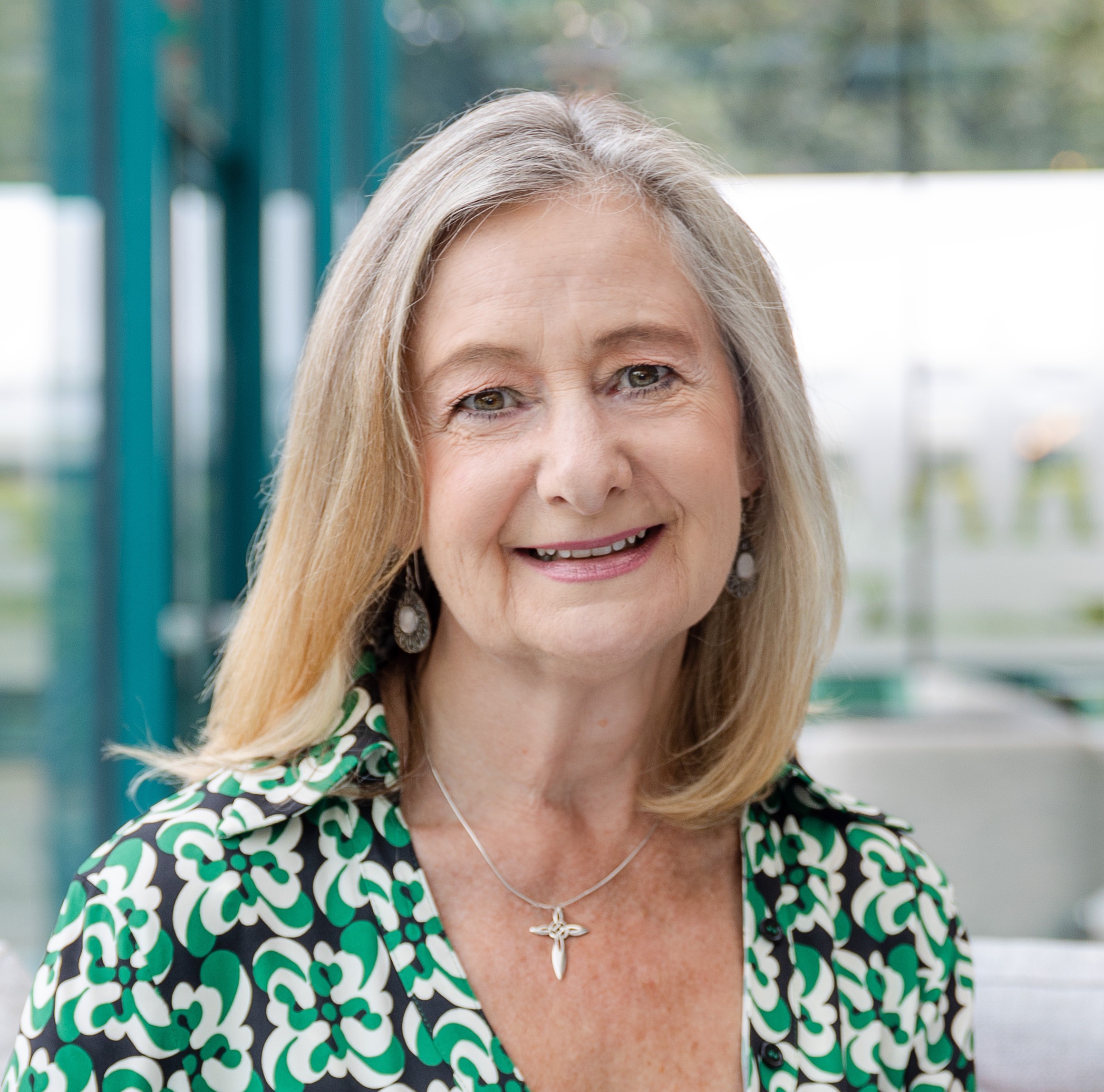 Smiling woman with shoulder-length blonde hair wearing a green-and-white patterned blouse and pendant necklace