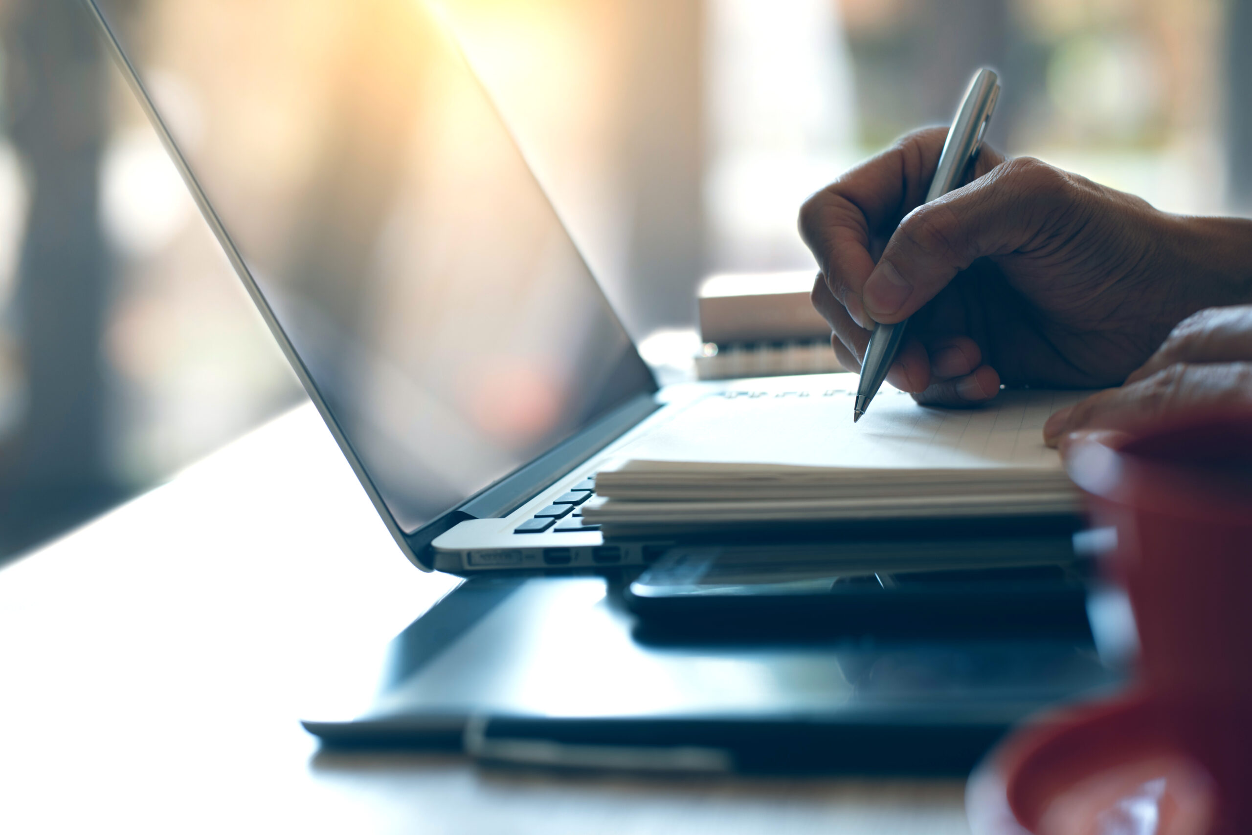Person writing notes beside an open laptop and stacked notebooks