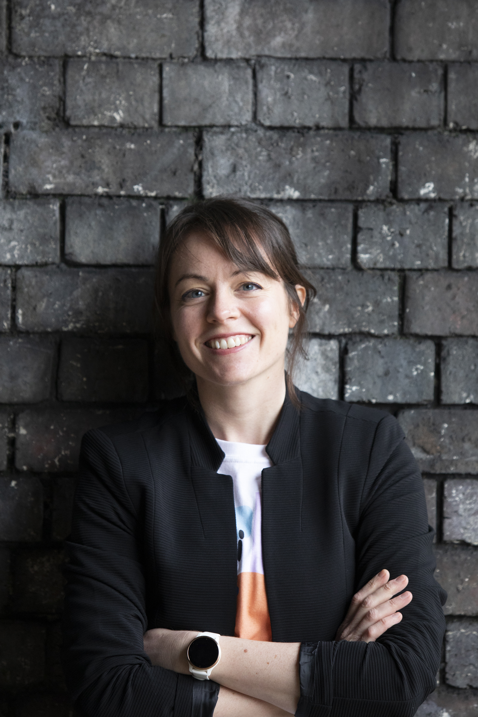 Smiling woman in a black blazer standing with folded arms against a dark brick wall