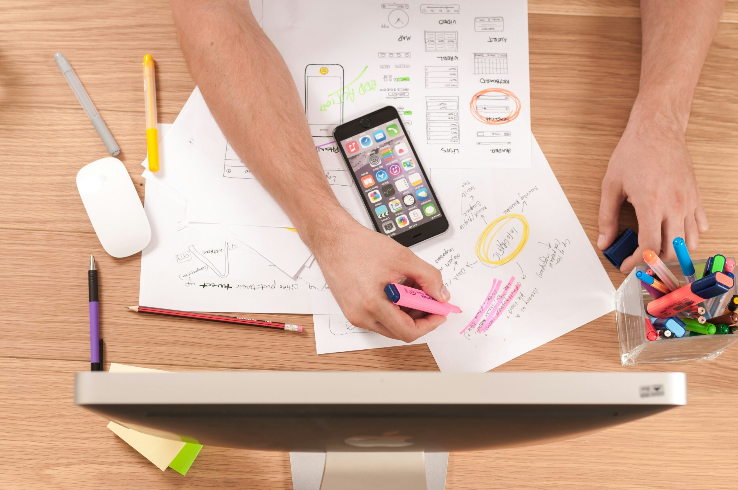People collaborate on website sketches and notes at a desk, with a smartphone, computer, pens and highlighters