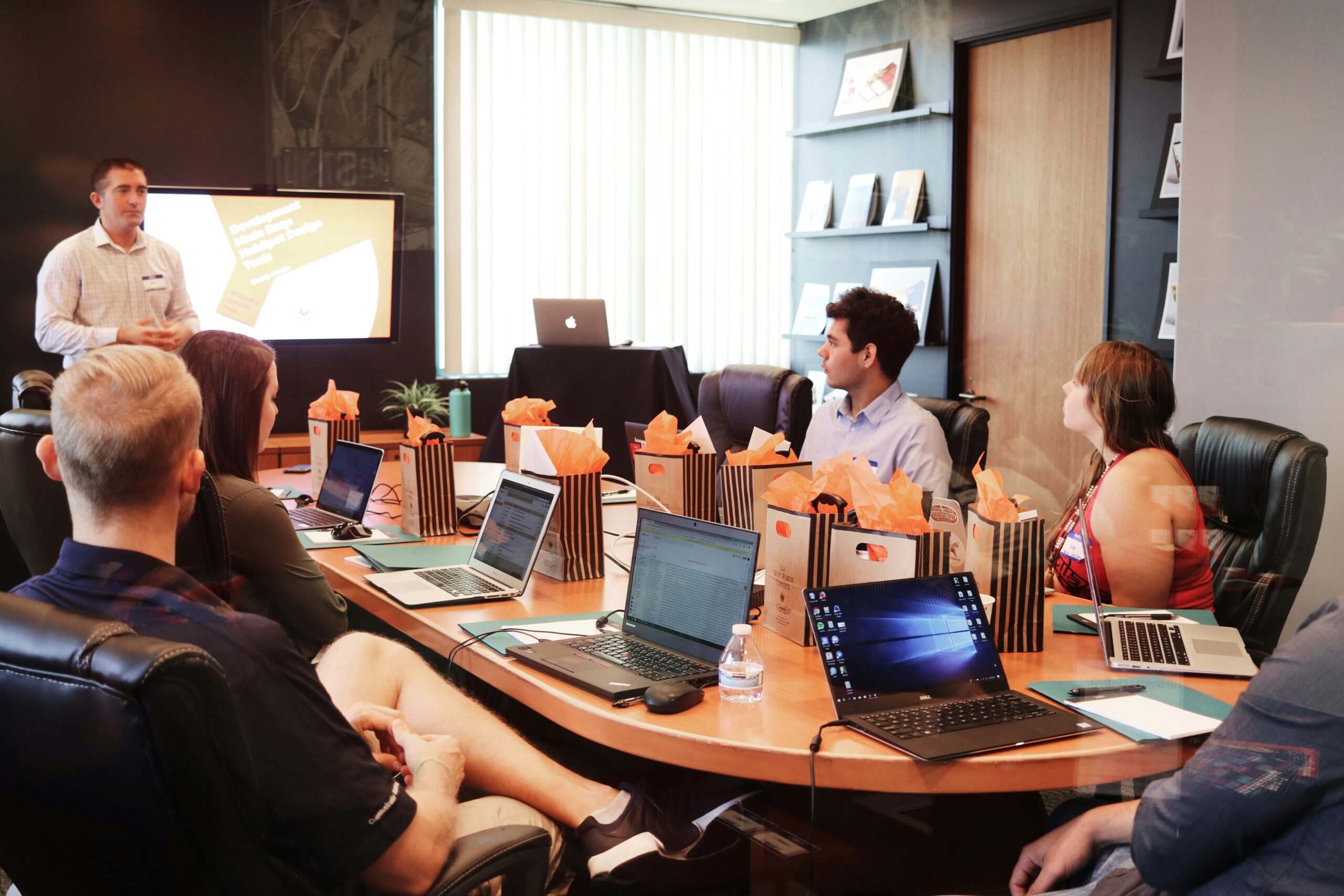 A presenter addresses a meeting of colleagues seated around a conference table with laptops and gift bags