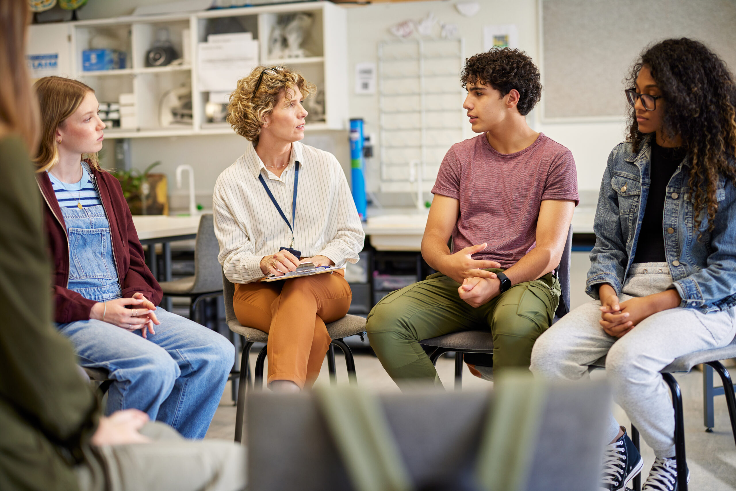 A teacher listens carefully to a boy speaking in a mixed group of students, addressing misogyny in schools through open discussion.