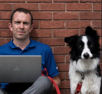 Man with a laptop sitting beside a black-and-white dog against a brick wall