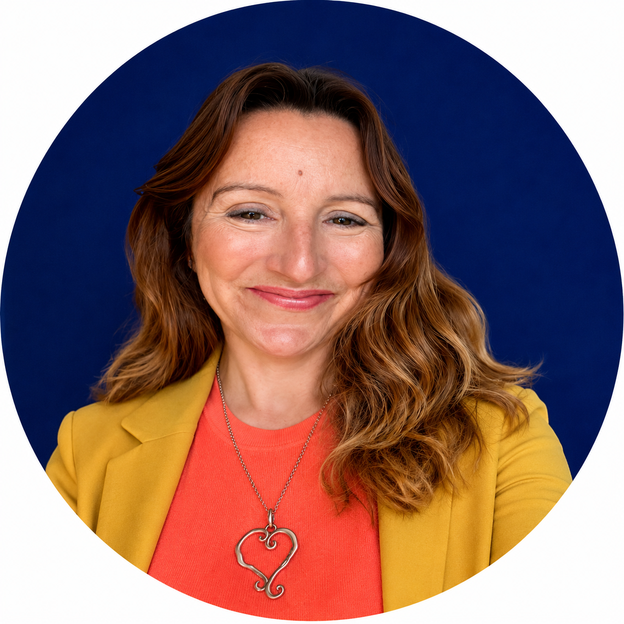 Smiling woman with long brown hair wearing a mustard blazer, coral top and heart-shaped pendant necklace