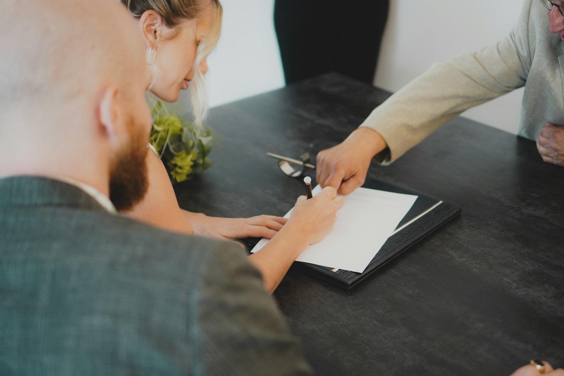 People signing a document at a table while another person points to the page